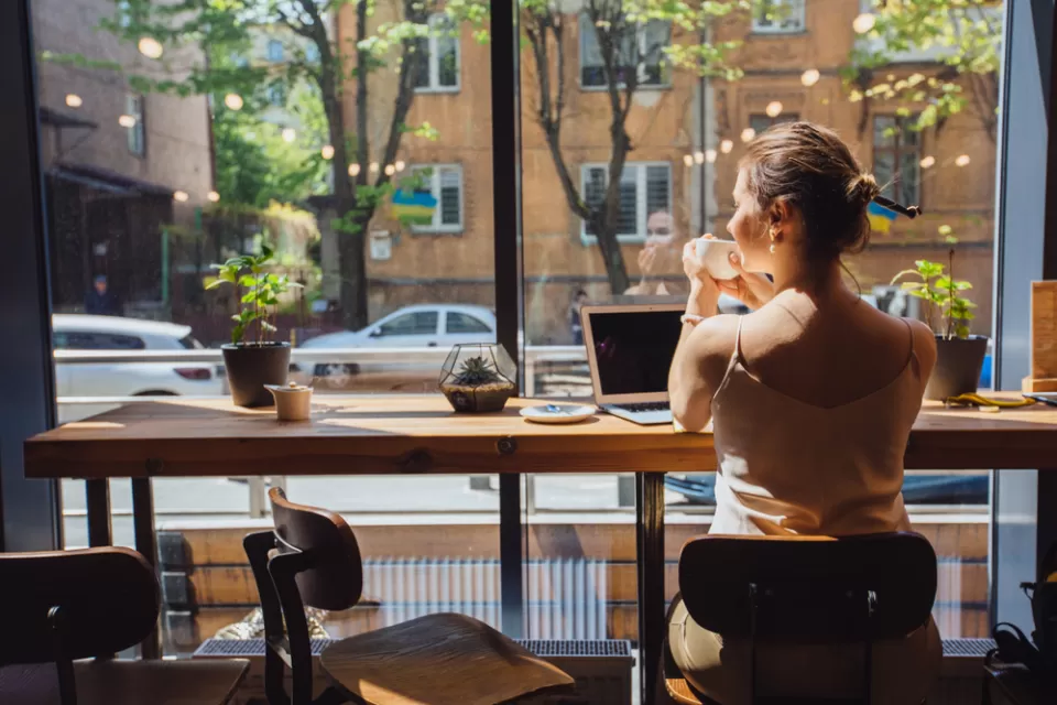 Woman sitting at cafe table with laptop, holding coffee cup and looking out at street
